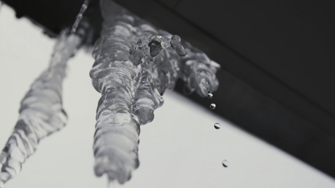Bottom view of melting icicle hanging from the roof in spring time. Stock footage. Water dripping from an icicle on grey sky background.